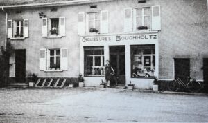 Une devanture de magasin portant l'enseigne peinte « Chaussures Bouchholtz » : deux vitrines garnies de souliers, une personne debout dans l'embrasure de la porte, et une bicyclette appuyée à droite.