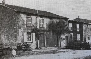 Un alignement de maisons de village à volets de bois, l'une portant un petit balcon de fer ; un tas de fagots est adossé au mur et le sol devant est en terre.