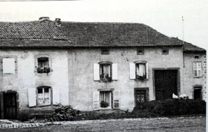 Une longue maison basse à toit de tuiles, volets ouverts et grande porte de grange ; un tas de bois de chauffage est posé devant, et des poules picorent près du seuil.