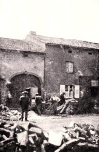 Des hommes débitent du bois à la scie devant une vieille ferme à porte cochère cintrée ; des tas de bûches occupent tout le premier plan.
