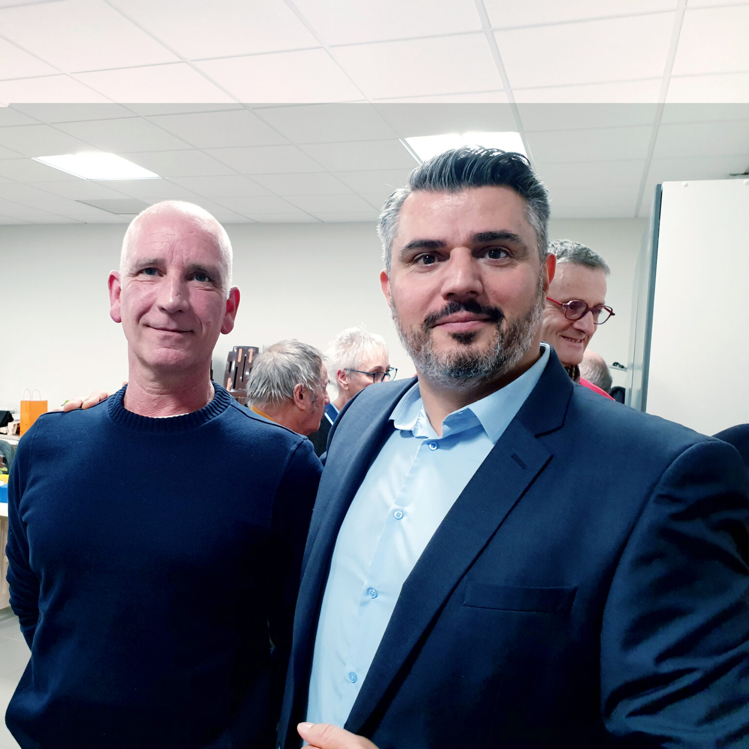 Deux hommes posent côte à côte, le bras sur l'épaule, lors d'une réception en salle ; d'autres invités discutent derrière eux.