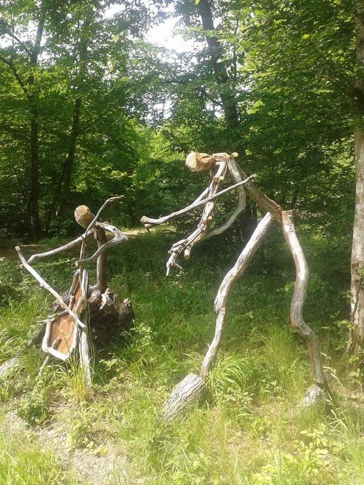 Une création de Land'Art en lisière de bois : deux silhouettes assemblées avec des branches et des rondins écorcés, l'une dressée sur de longues jambes, l'autre accroupie sur une souche, au milieu de l'herbe haute.